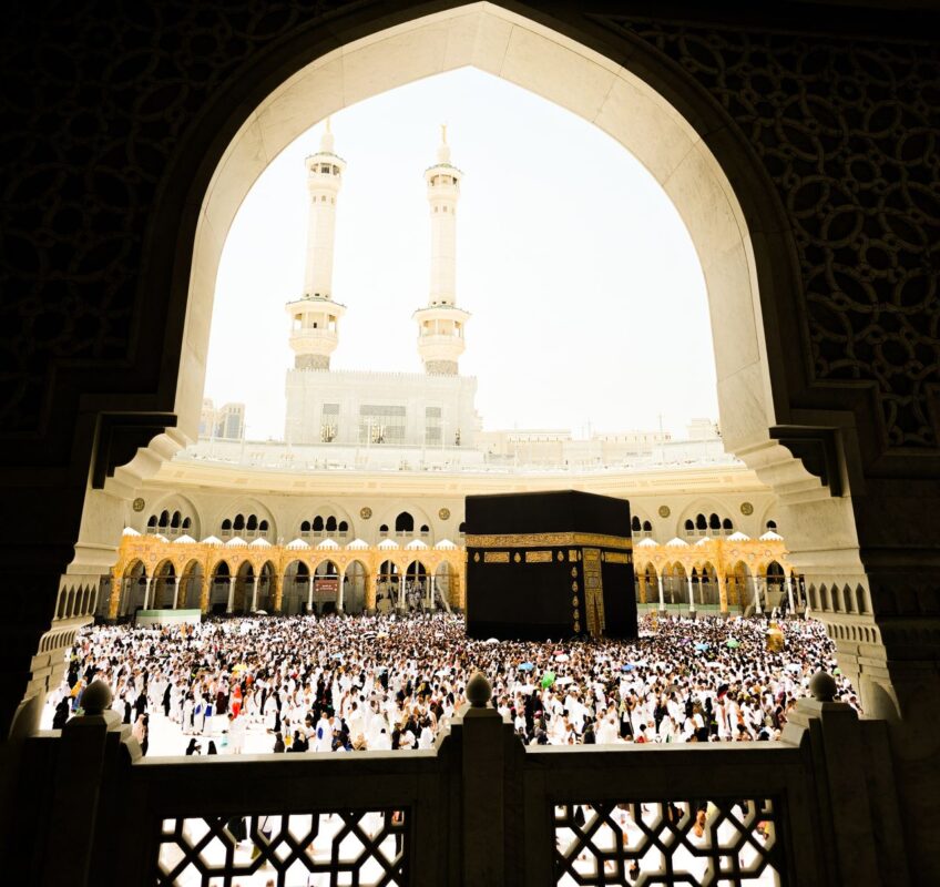 A large crowd of pilgrims surrounding the Kaaba during daylight, with a view of the Grand Mosque's architecture.